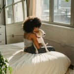 A woman with afro hair lovingly holds a dog in a sunlit bedroom, symbolizing pet companionship.