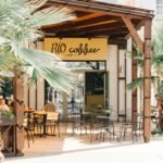 An inviting outdoor café under palm trees with wooden tables and chairs on a sunny day.
